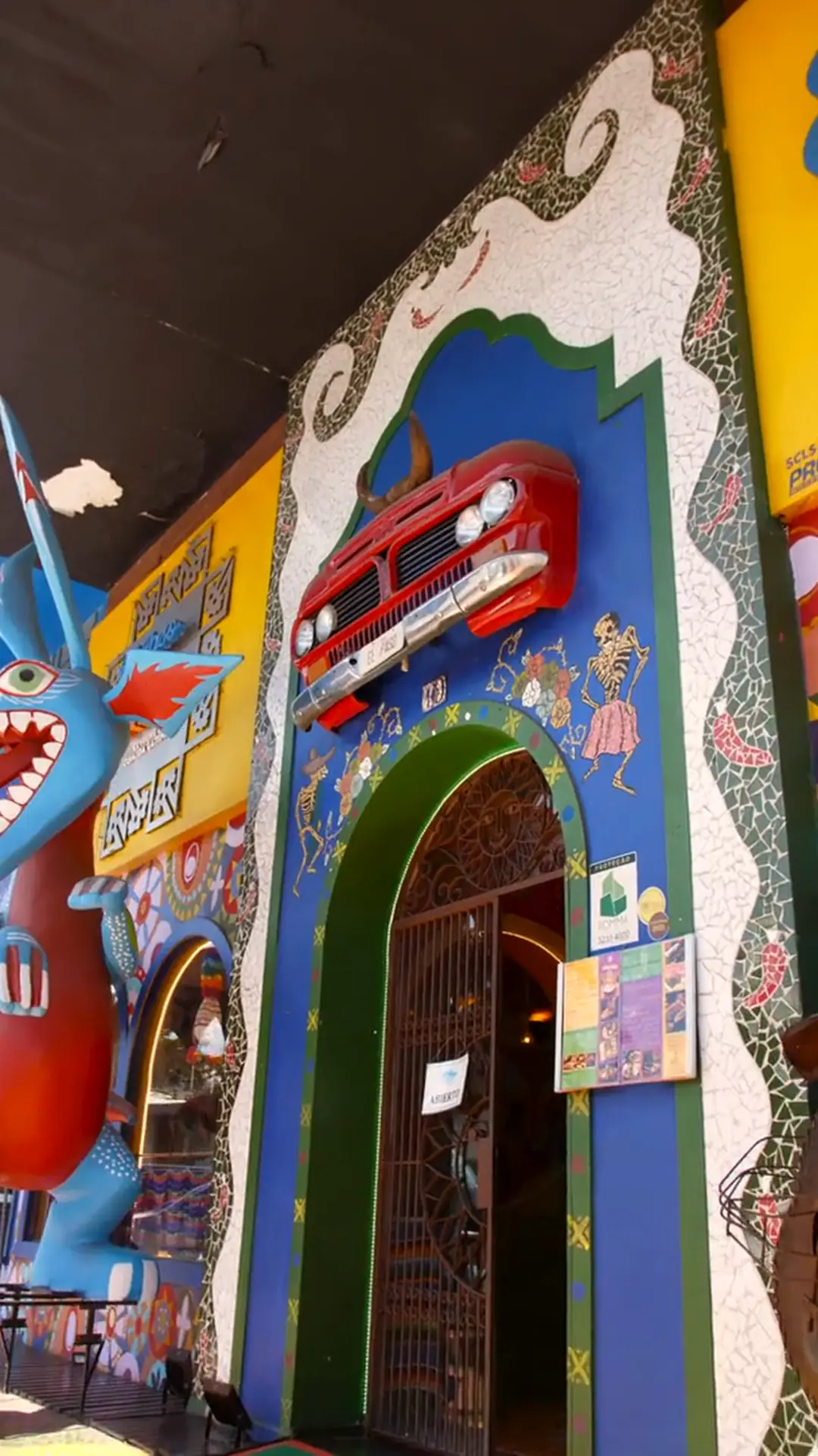 Fachada do El Paso: mural colorido com alebrije e um carro vermelho preso à parede, sobre o arco verde da entrada