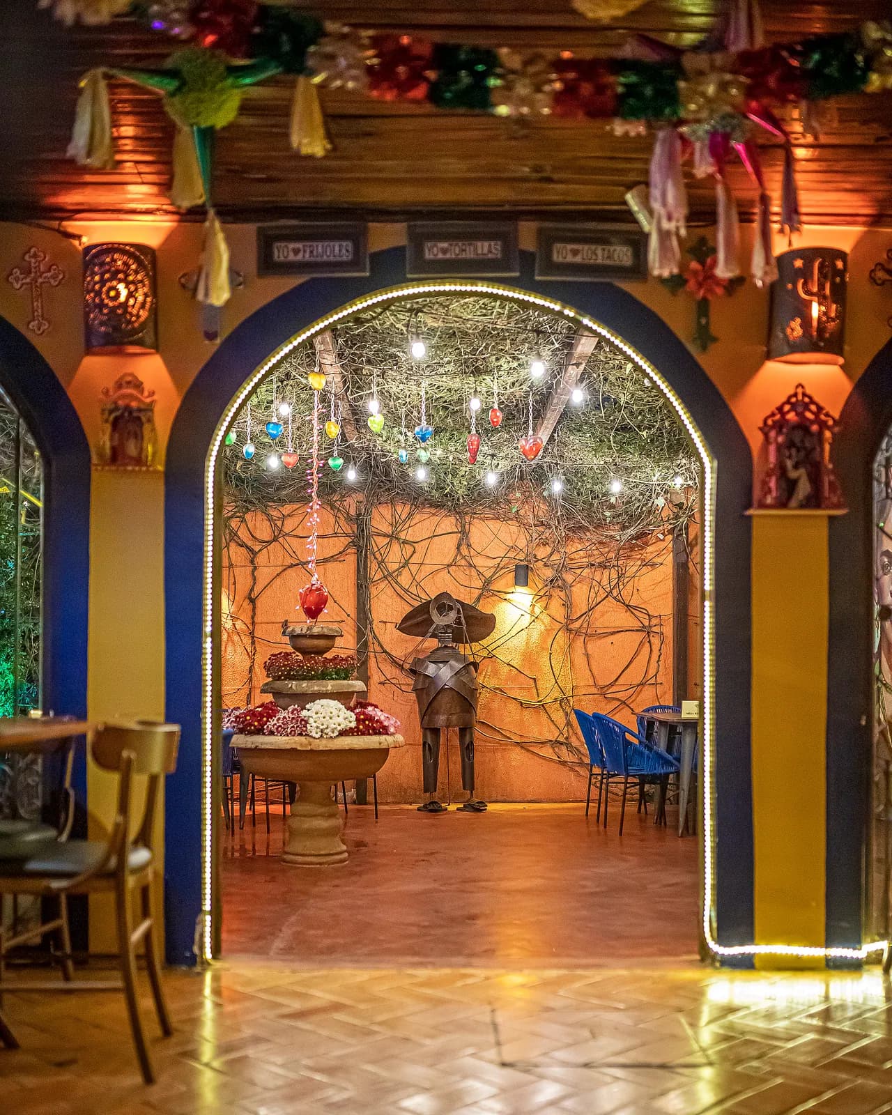 Arco decorado com placas "Yo amo tacos" emoldurando o pátio com fonte e escultura de mariachi