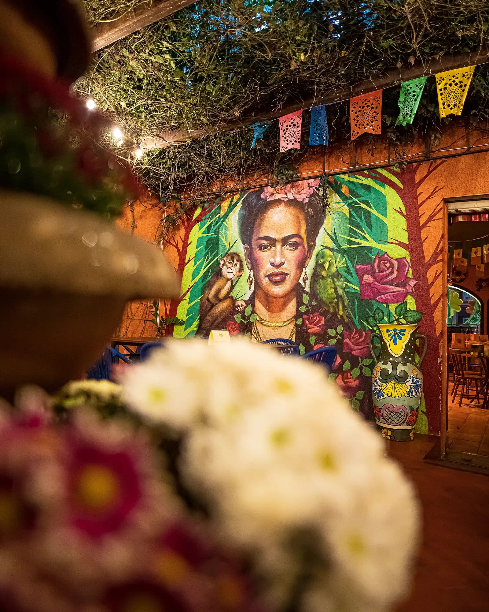 Mural da Frida Kahlo no pátio, com flores em primeiro plano e papel picado